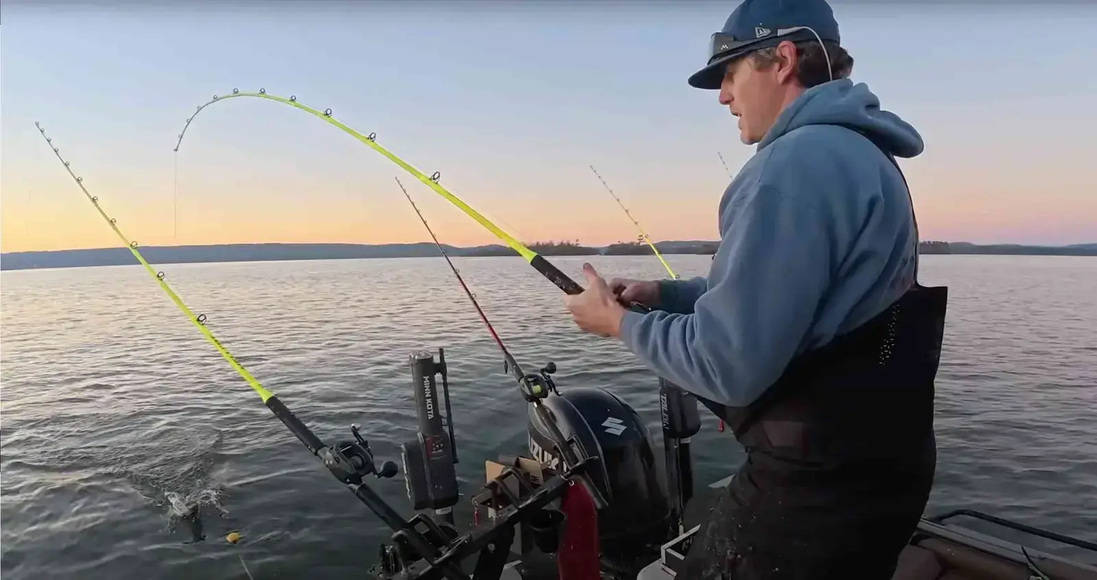 An angler battles a catfish on a neon Hellcat rod at twilight, with the fish’s swirl visible by the boat.